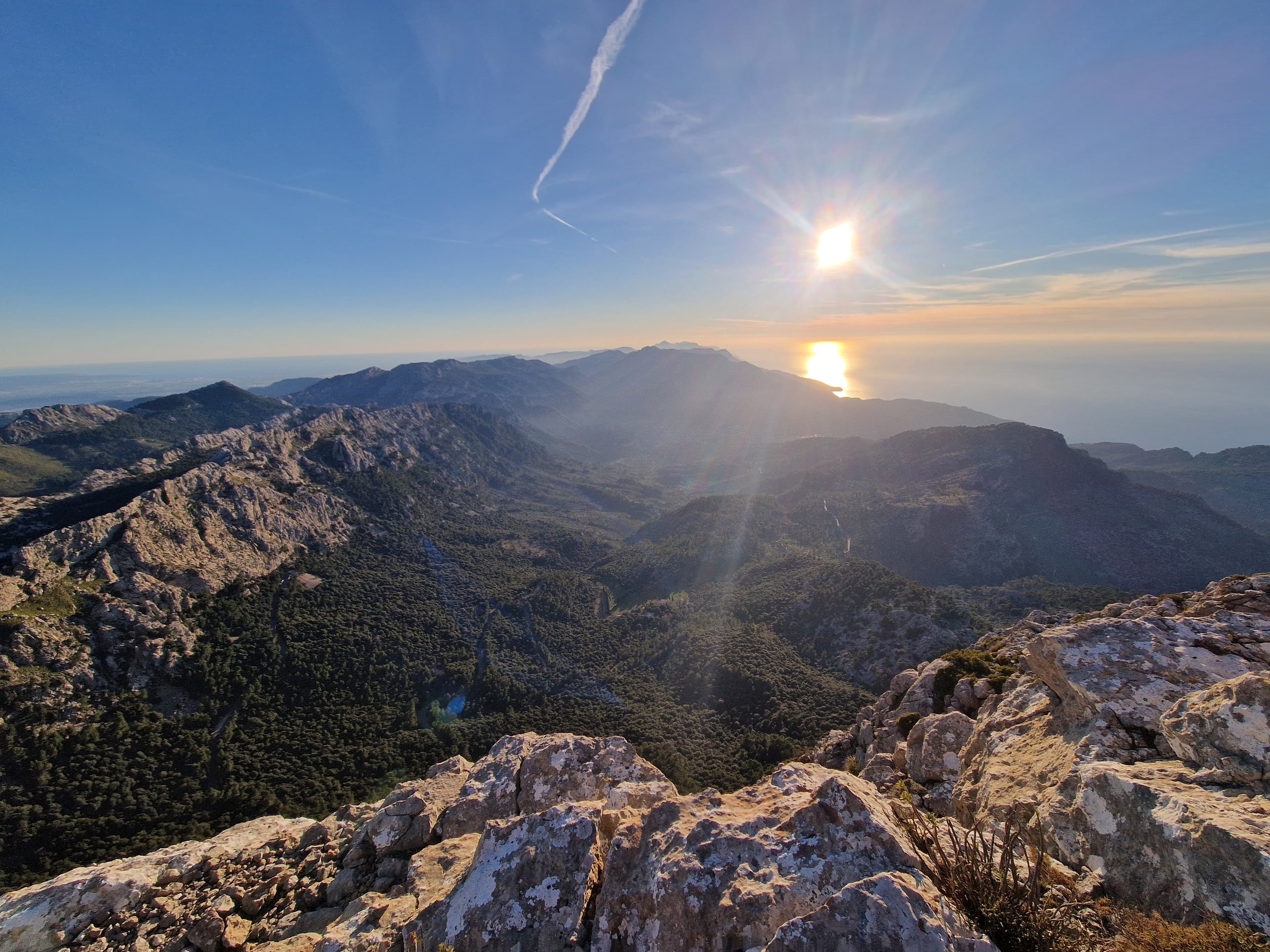 Sunlight over a rugged mountain range and forested valley, viewed from a rocky summit.