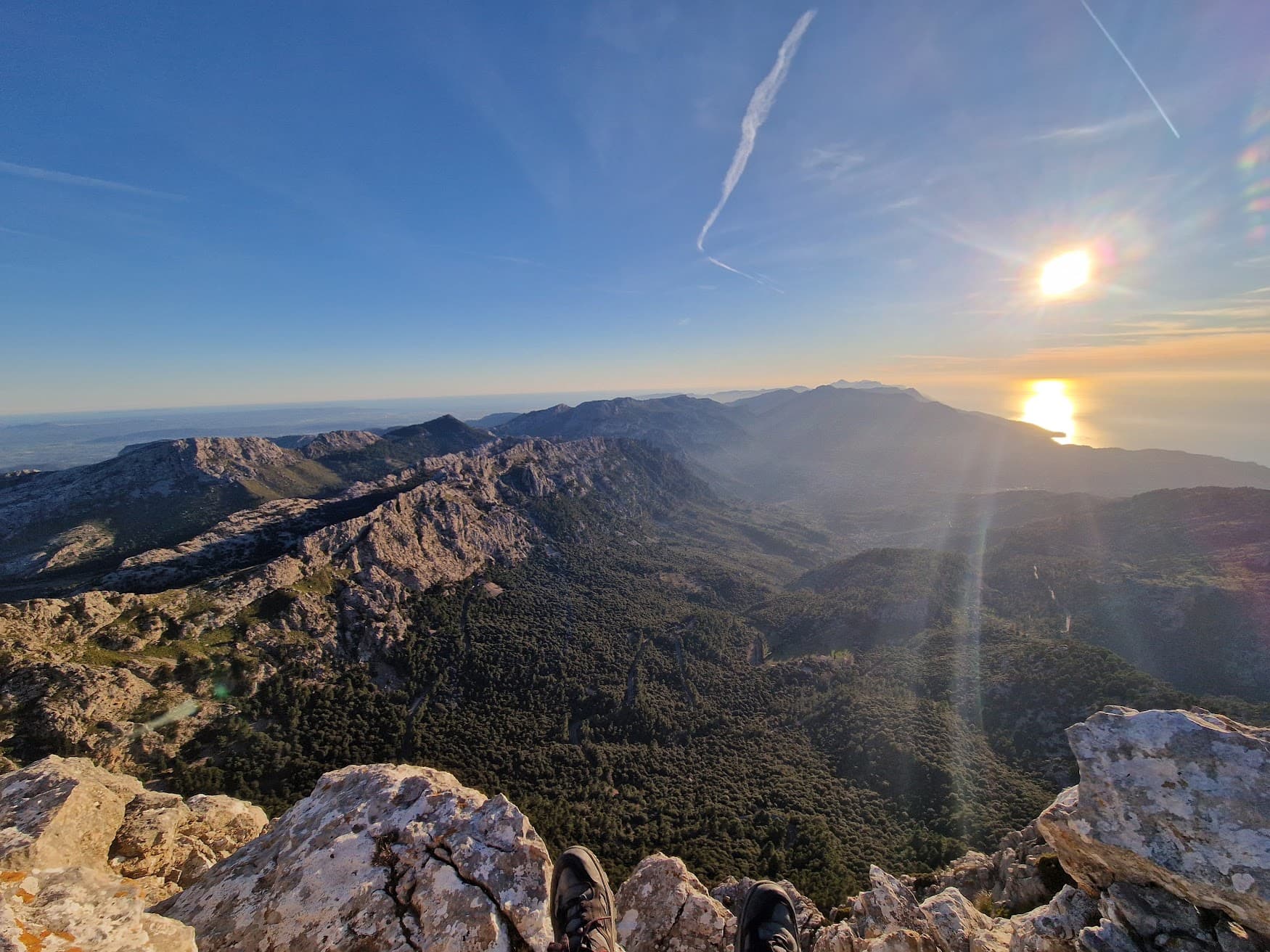 Hiking boots on a rocky summit overlooking a vast forested valley and coastal sunset.