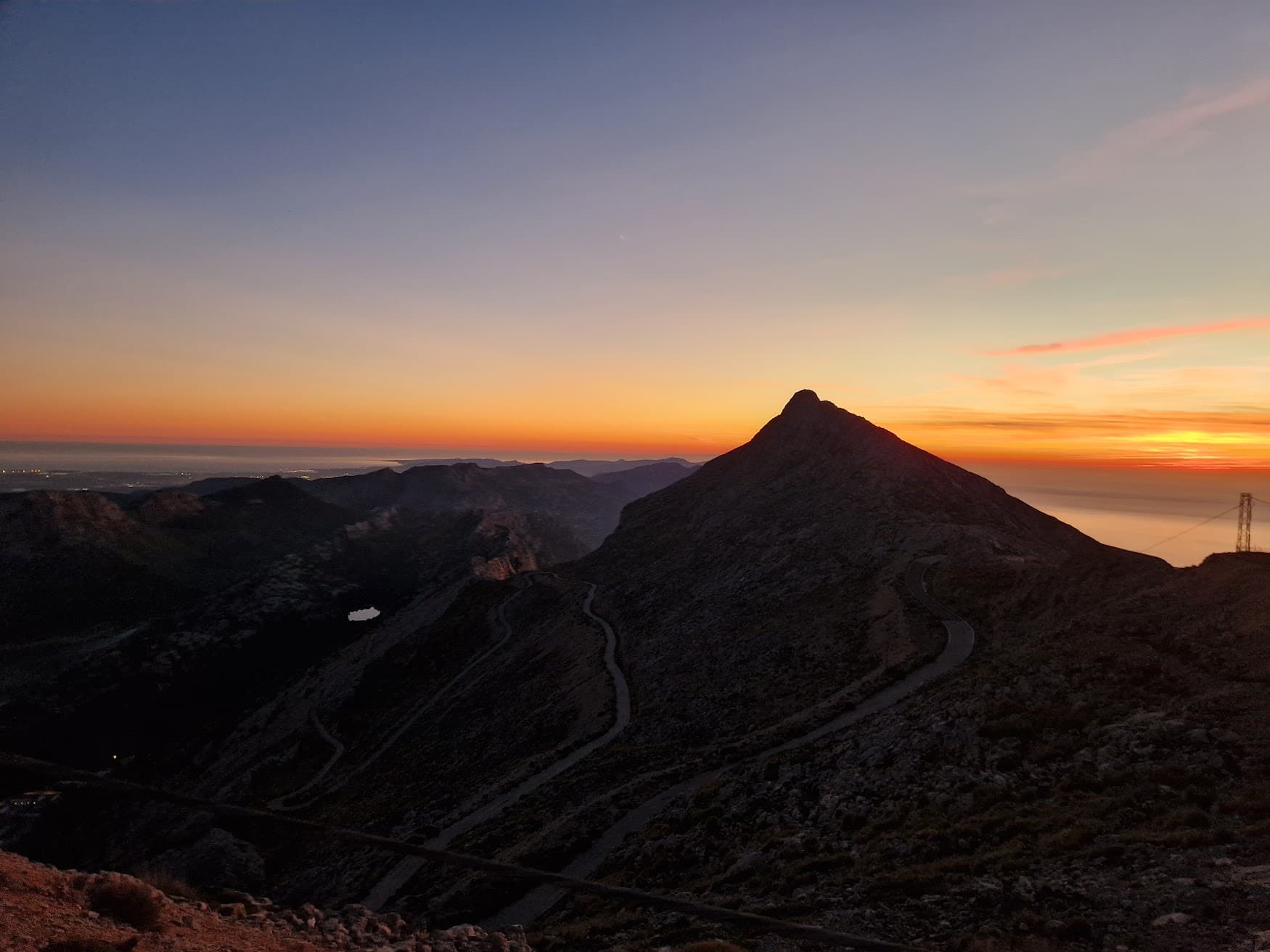 Winding road snakes up a dark mountain peak against a vibrant orange and blue sunset.