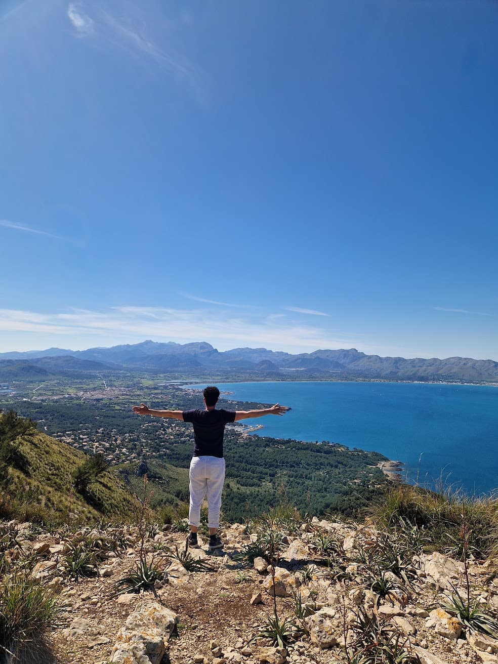 Man with arms wide on a mountain peak overlooking a scenic bay and mountains.