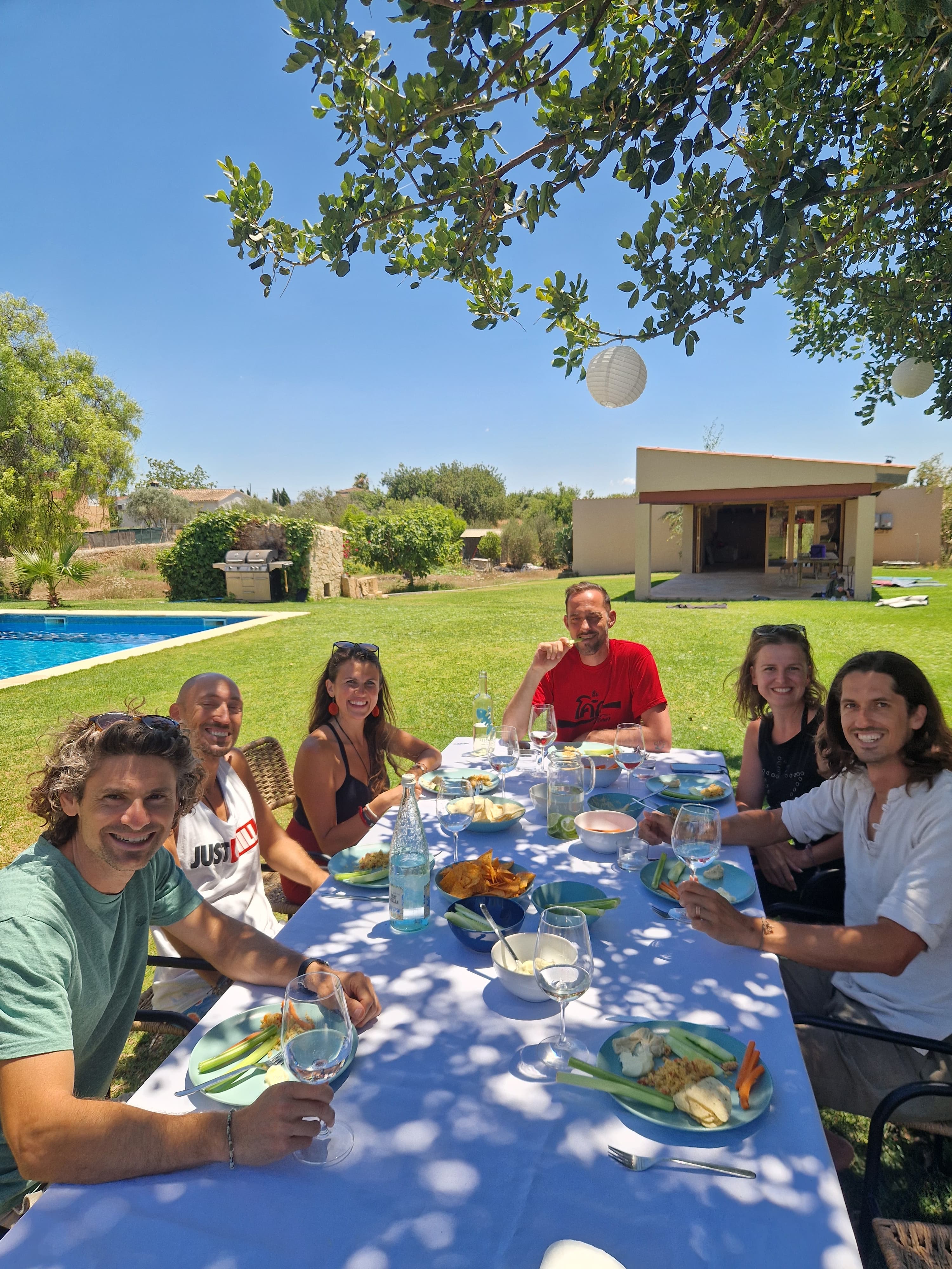 Six friends smiling around an outdoor dining table in a sunny backyard with a pool.
