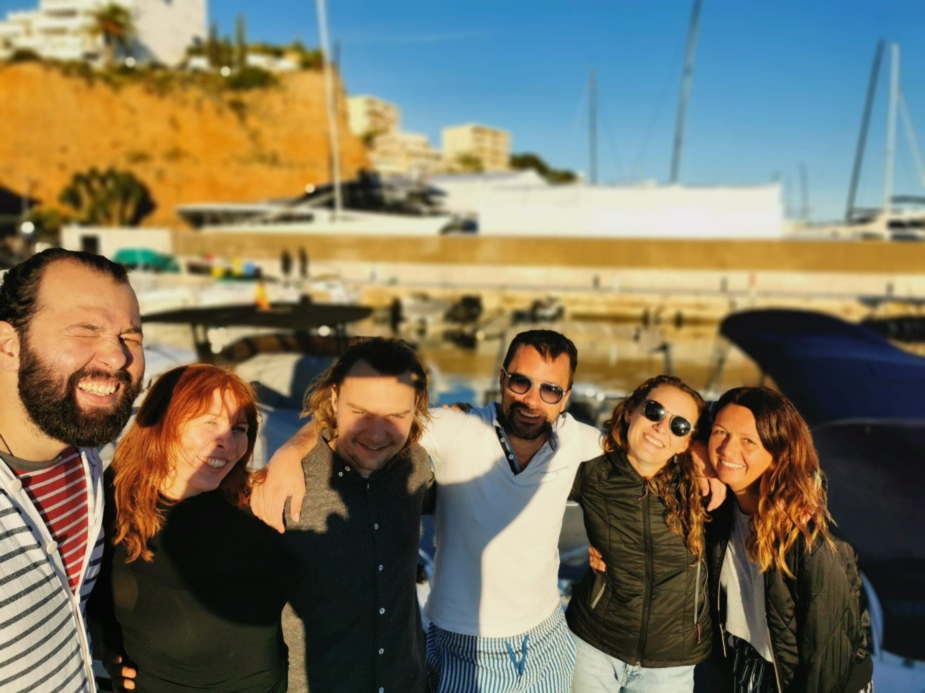 Six smiling friends pose at a sunny marina with boats and a hillside background.