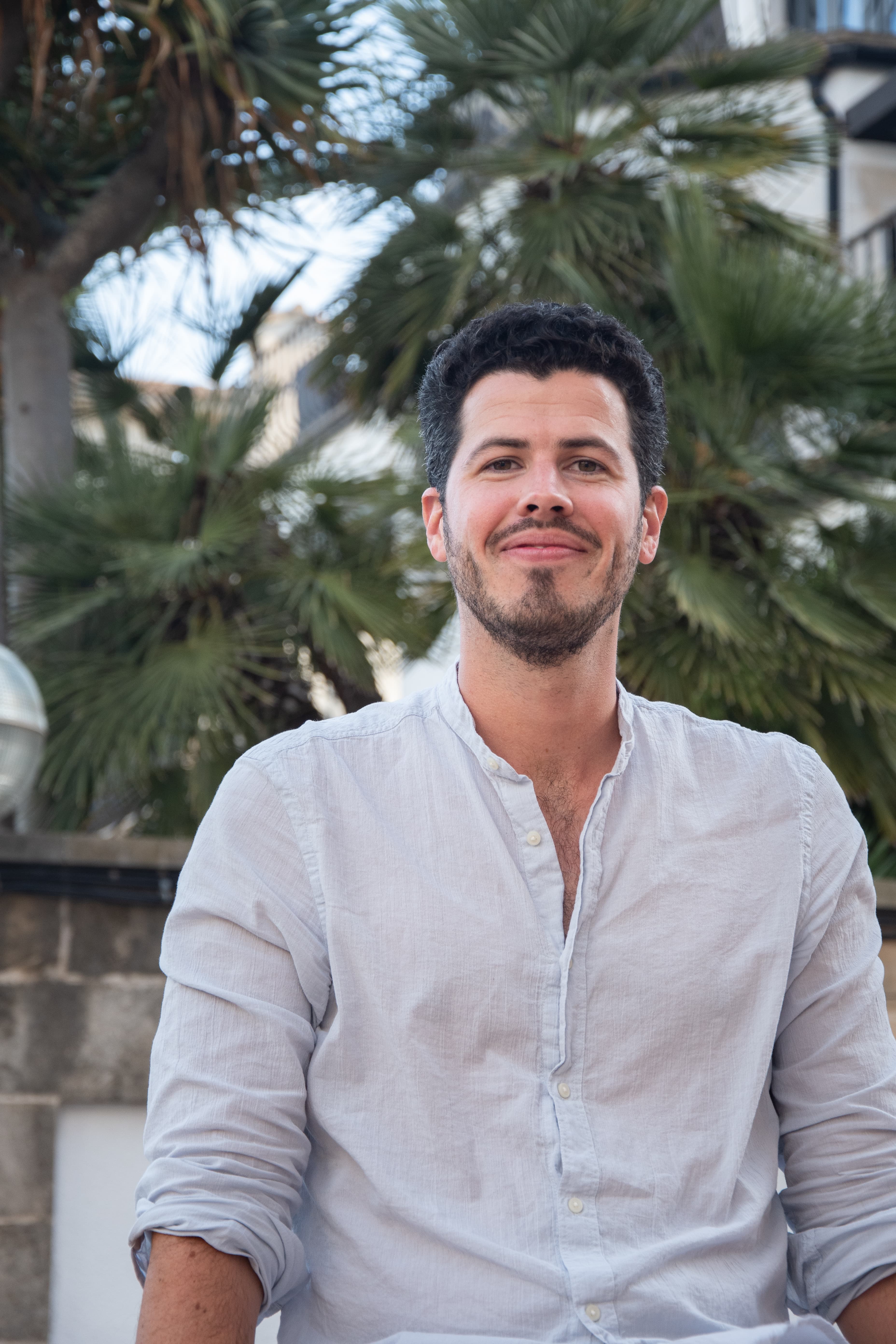 Smiling man with dark curly hair and beard in a light blue linen shirt outdoors.