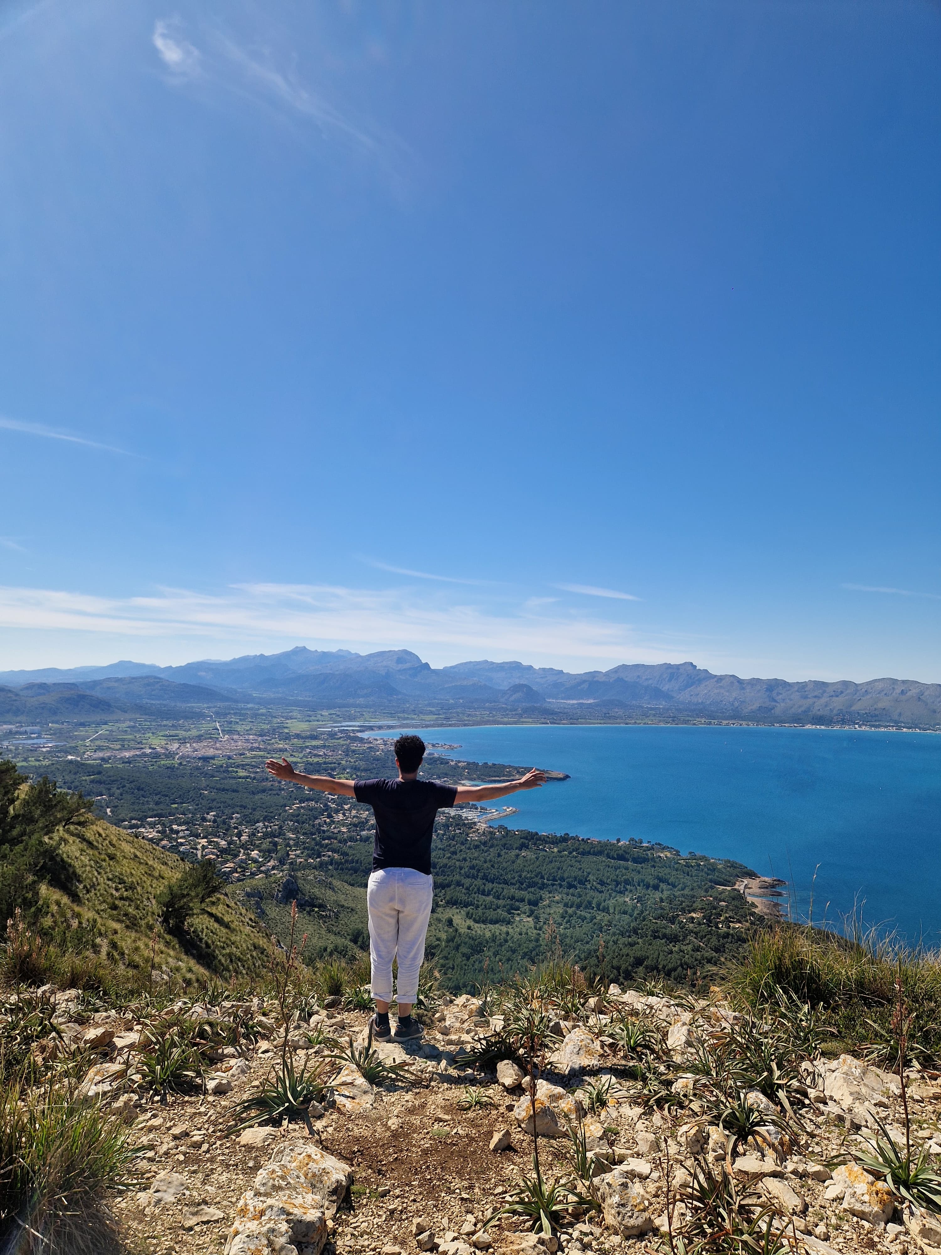 Man with arms outstretched on a rocky peak overlooking a blue bay and mountains.